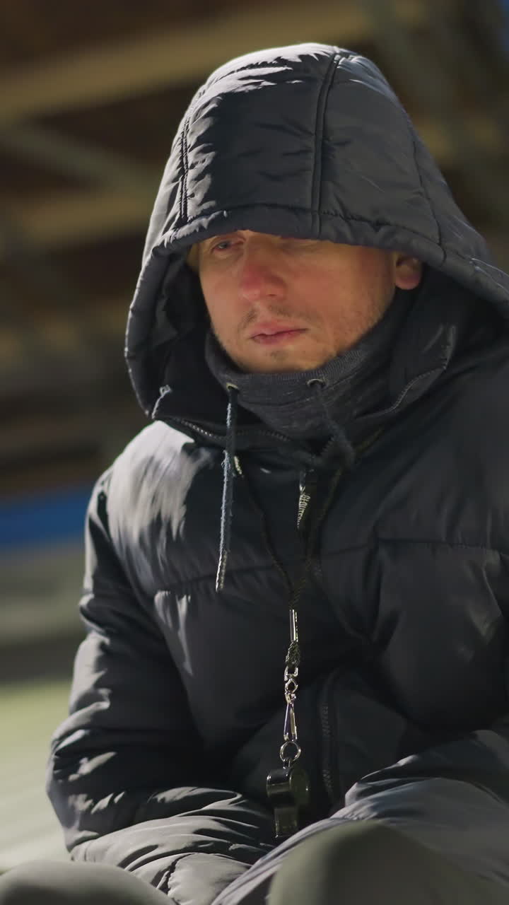 un hombre con un traje negro y una chaqueta con capucha se sienta solo con las manos entre las piernas, pareciendo sobrio, está en un estadio con un fondo débilmente iluminado