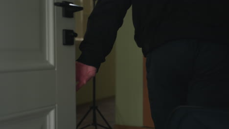 man seen from waist down holding travel bag handle walking into apartment through open door, wearing black jacket and pants, smooth floor under soft daylight