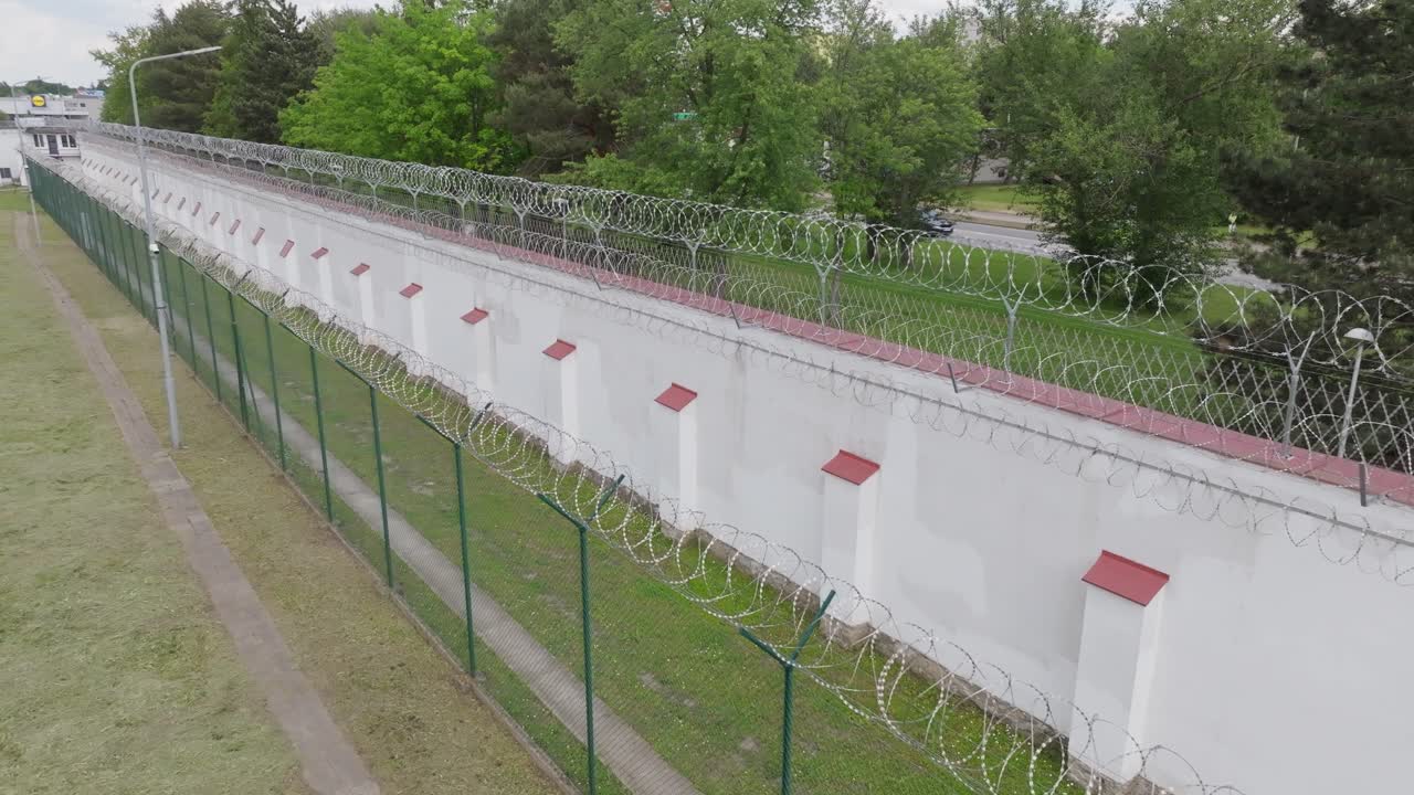 Elevated drone tracking shot moving along long stretch of fenced prison wall with security systems visible