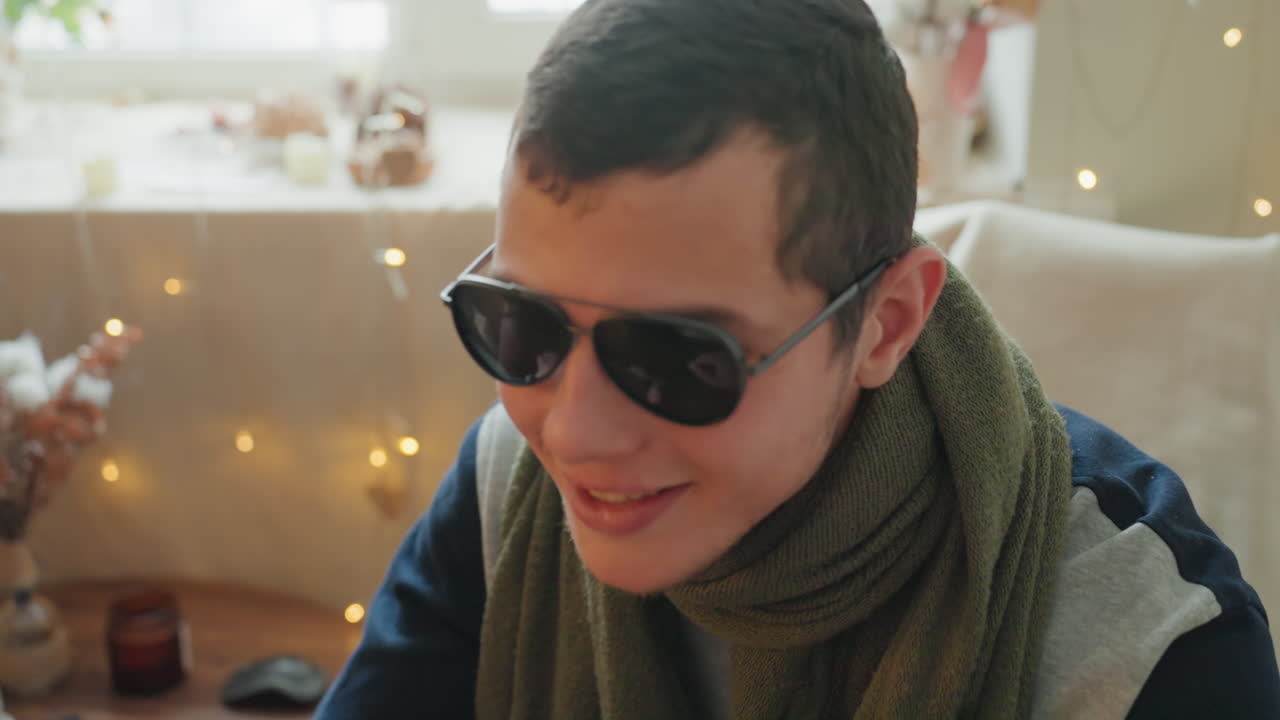 White man dons sunglasses near window. Cozy living scene shows playful transformation from focused student to confident style persona scarf, laptop, warm fairy lights and dried flowers create