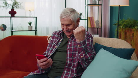 Overjoyed-senior-grandfather-man-hold-smartphone,-excited-about-mobile-app-sport-bet-bid-win-at-home