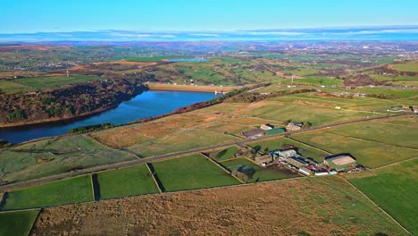 Imágenes-Aéreas-De-Drones-Del-Tráfico-En-Una-Carretera-Rural-Con-La-Autopista-M62-A-Lo-Lejos,-Cruzando-El-Agua-De-Scammonden-Con-El-Lago-De-Tierras-De-Cultivo,-El-Embalse-Y-Las-Amplias-Vistas-Abiertas-De-Las-Colinas-Peninas