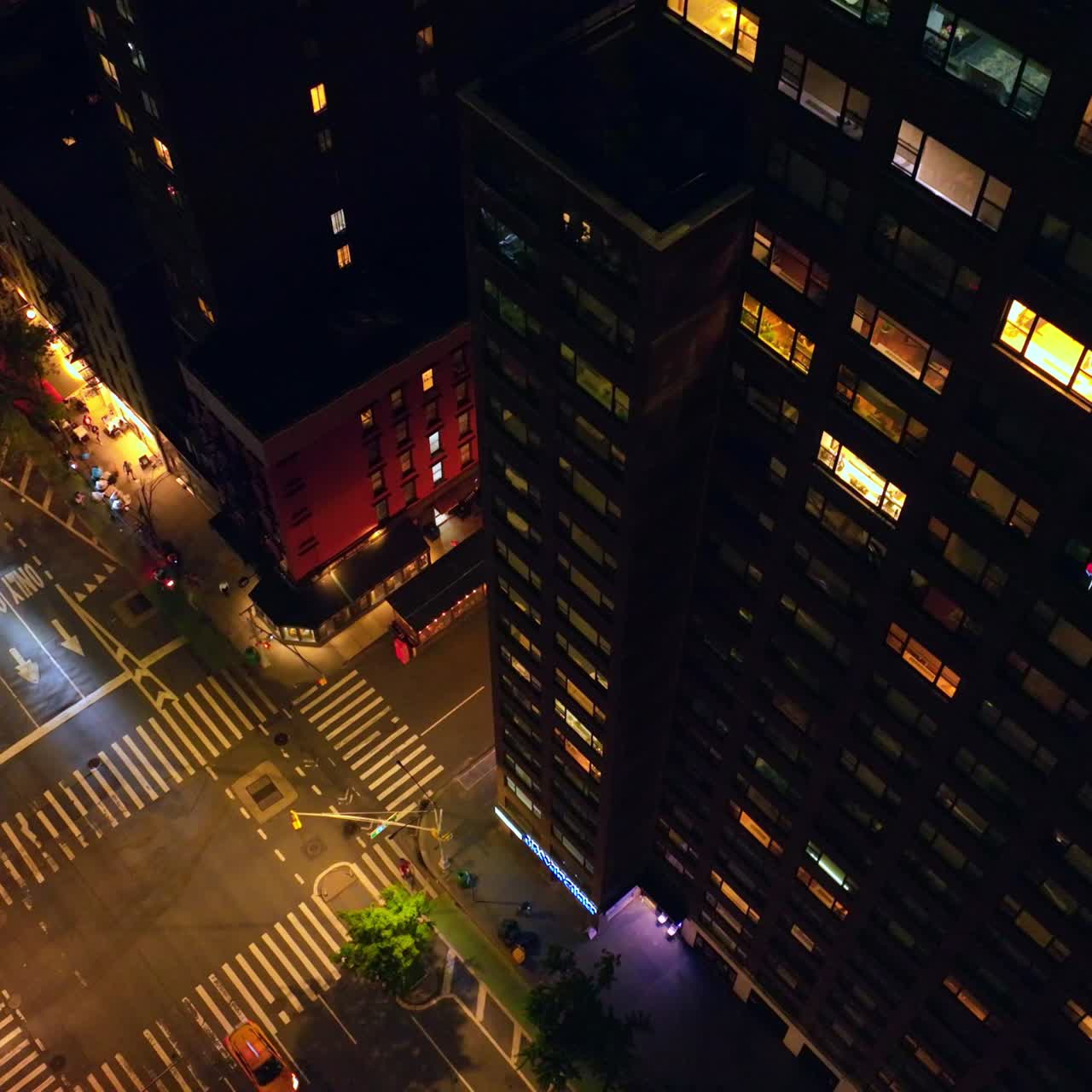 Dark multi-storied buildings with lit windows at night. Drone descending over the well-lit roads with busy traffic