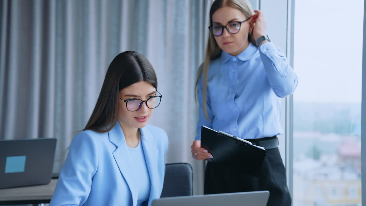 Female office colleagues having conversation while looking at the screen of laptop. Busy young ladies in glasses discussing work issues.
