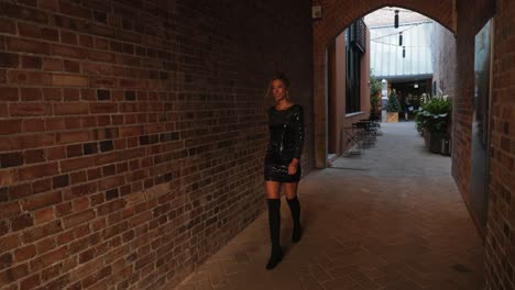 Confident-attractive-woman-in-tight-black-dress-and-knee-length-boots-walks-down-brick-alley-towards-camera,-turns,-smiles