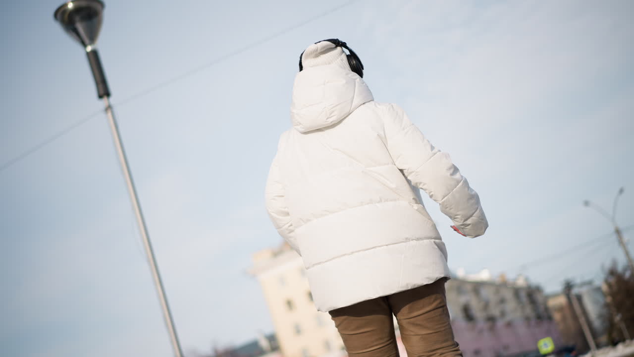Back view of creative dancer swirling arms under tall street pole on urban plaza, clad in white winter jacket and tan pants, moving with rhythm against backdrop of city buildings and winter sidewalk