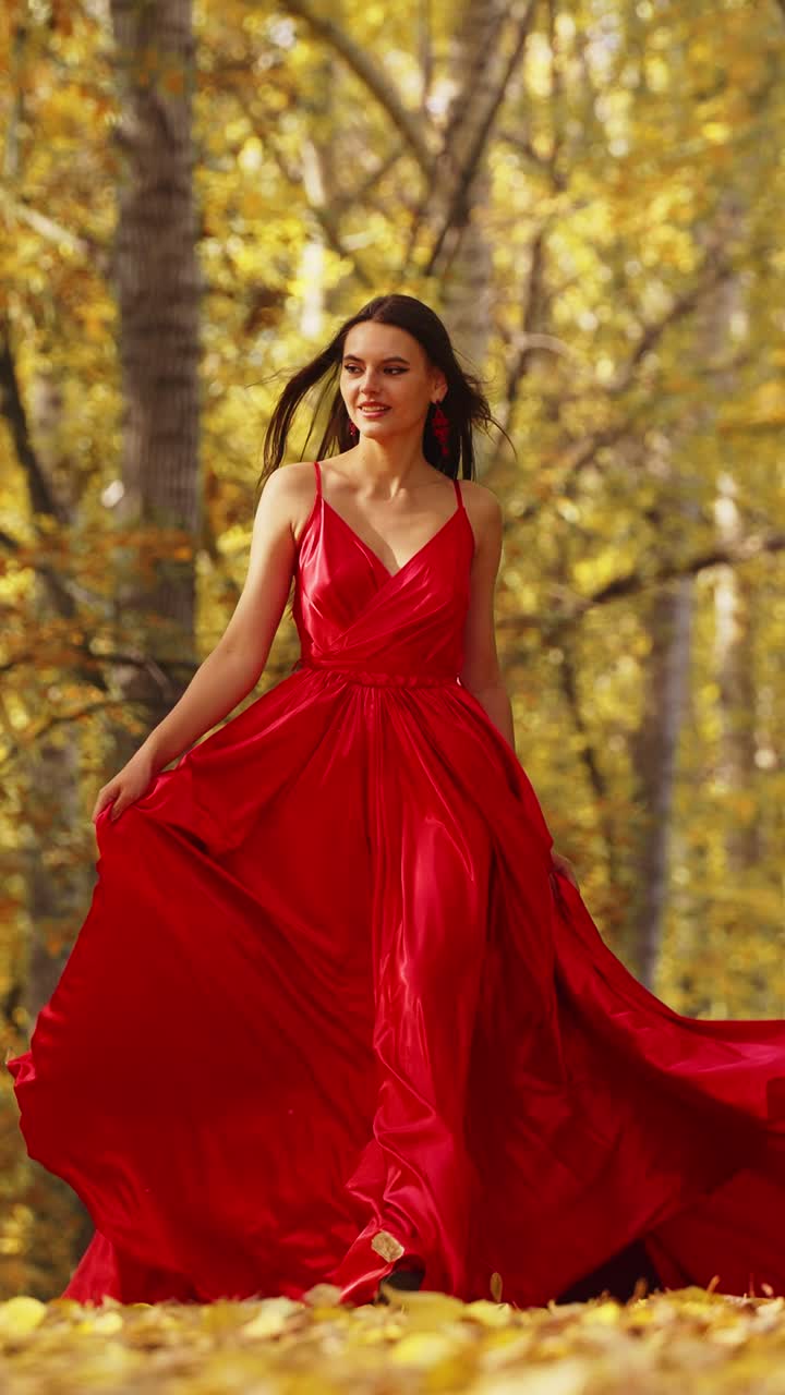 Una donna elegante con un meraviglioso abito rosso cammina con sicurezza in una serena foresta autunnale, circondata da vivaci foglie dorate, mostrando eleganza e bellezza nella natura.