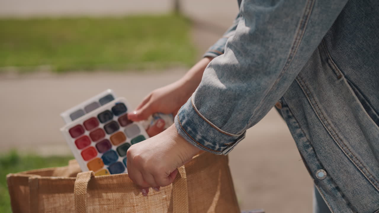 Artista che ripone la tavolozza degli acquerelli nella borsa vicino a un sentiero, giacca di jeans e borsa di iuta nell'inquadratura, le mani fanno scivolare la tavolozza compatta in un contenitore riutilizzabile, sistemando pennelli e forniture per un tragitto casual.