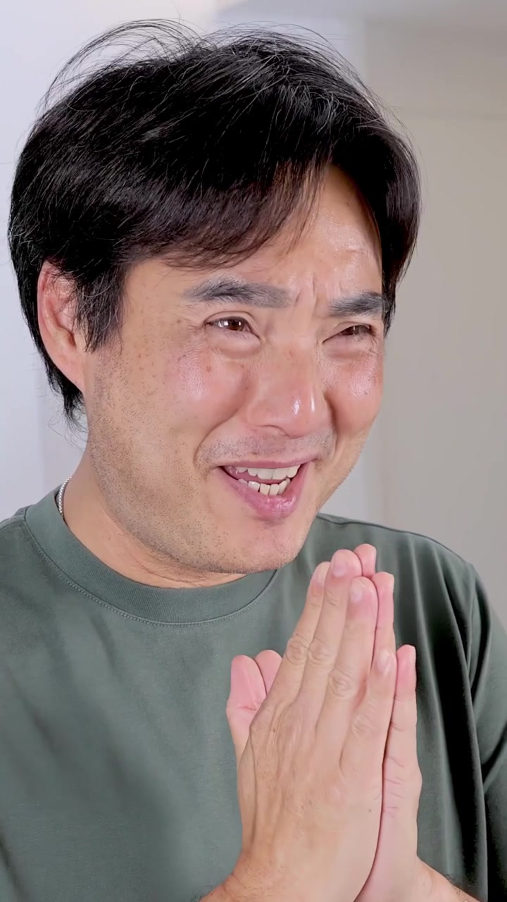 明るく照らされた屋内で、伝統的な敬意を表す挨拶のジェスチャーを行い、暖かく微笑むアジア人男性のミディアムクローズアップ。