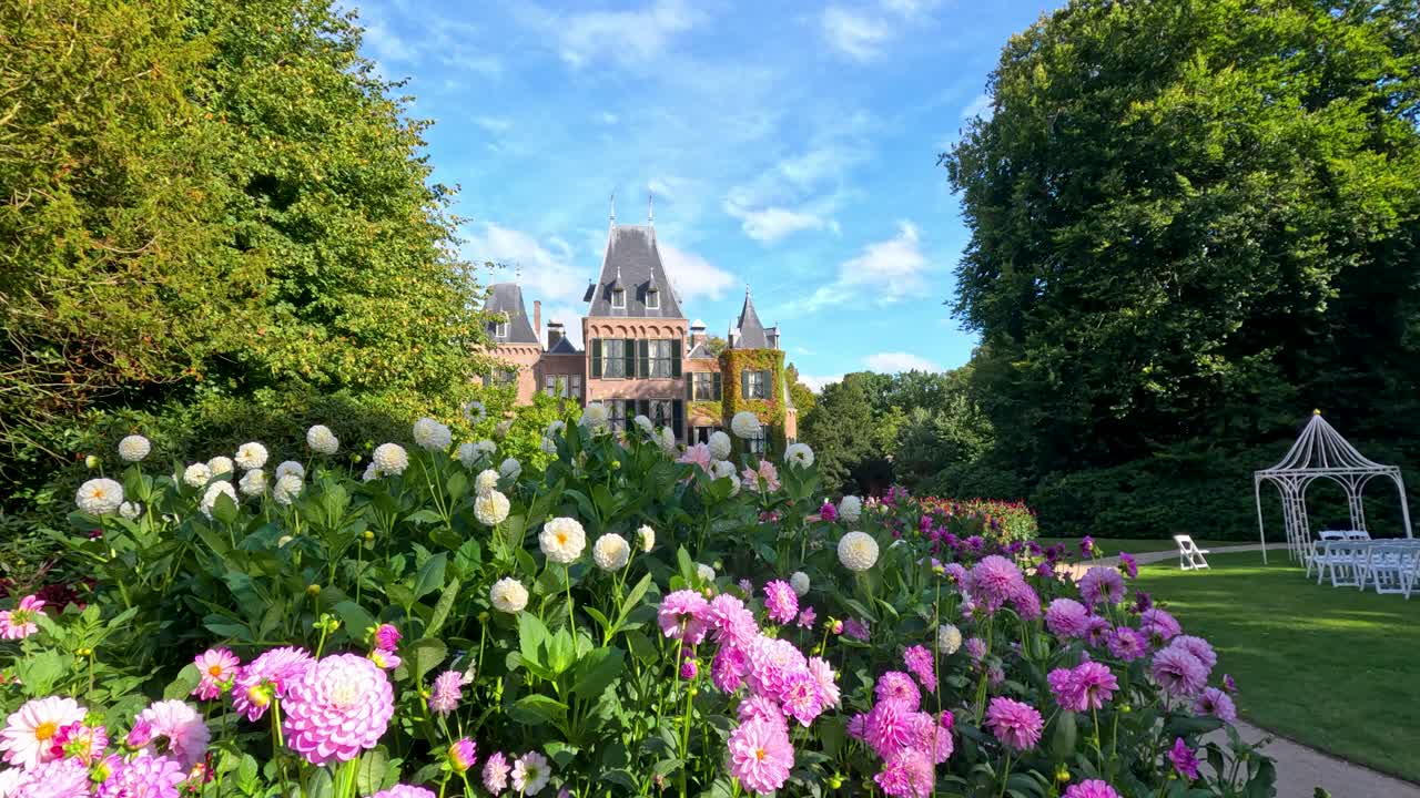Camera glides through vibrant dahlia garden toward a grand castle, under bright daylight with lush greenery and blue sky in Lisse, Netherlands