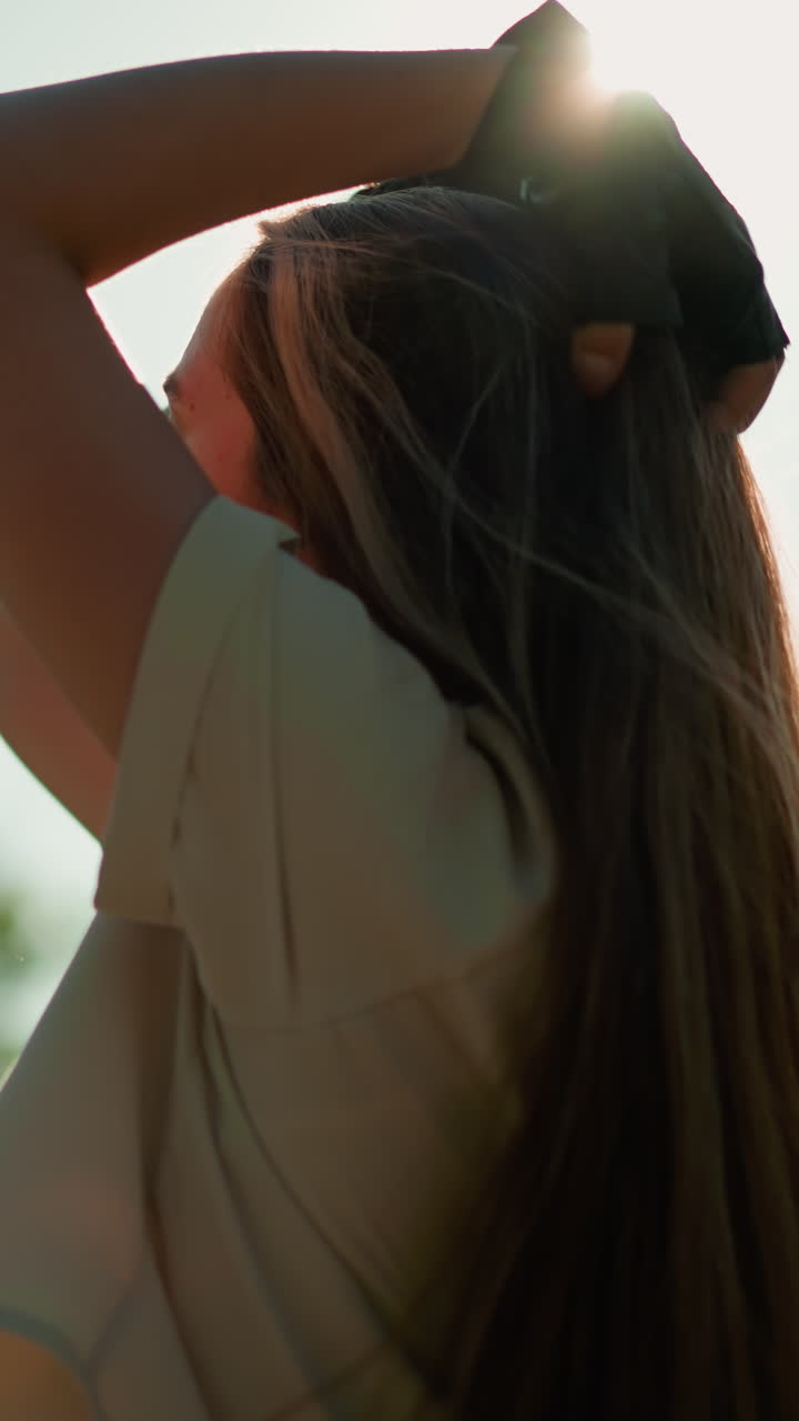 검은 장갑을 입은 여자가 머리를 뒤로 기울여 머리를 조정하면서 빛이 부드럽게 반사되는 동안, 조용한 초록색과 흐릿한 나무로 둘러싸여 있습니다.