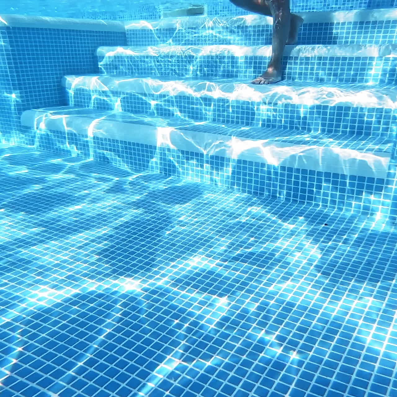 Transparent underwater background in the swimming pool. Underwater view of a boy's body coming down into the pool on stairs and swimming in blue clear water.