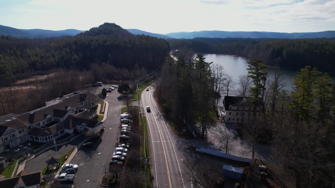 gran instalación médica y casa colonial cerca de un lago y coches de crucero en una carretera con cielo azul y aguas tranquilas entre bosques de hoja perenne y colinas onduladas