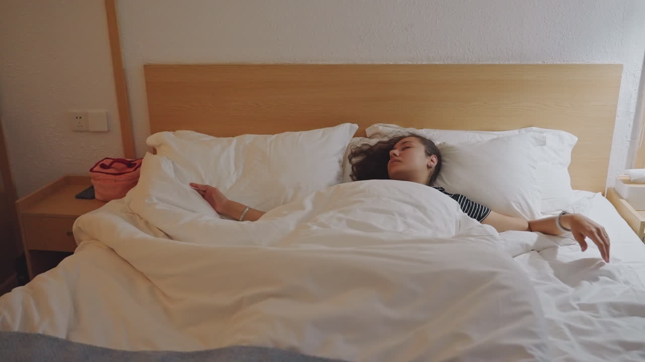 Behagelig hotelmorgen. Fredelig morgen, hvor en gæst strækker sig i hotelsengen under blødt daggrylys. Roligt byophold med en gæst, der afslappet strækker sig på den bløde seng i det tidlige morgenlys.