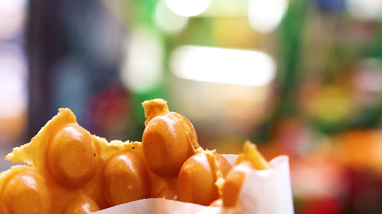 Close-up of hands reaching for bubble waffles in a paper bag with a colorful blurred background.