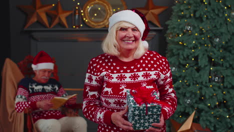 Senior-grandmother-in-festive-sweater-presenting-Christmas-gift-box,-smiling,-looking-at-camera