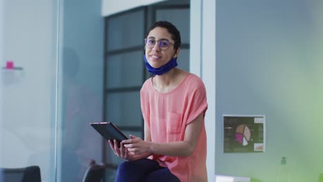 Portrait-of-caucasian-woman-with-lowered-face-mask-smiling-while-using-digital-tablet-at-modern-offi