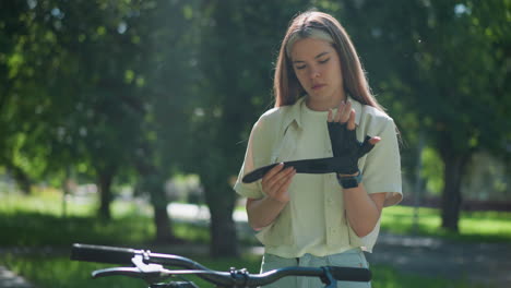 young woman standing beside bicycle, carefully adjusting biker glove with focused expression, as sunlight filters gently through trees in blurred background