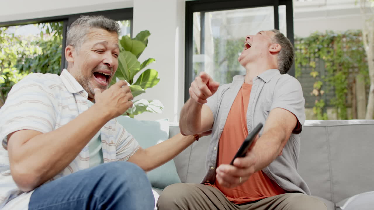 Laughing together, senior friends sitting on couch and holding smartphone