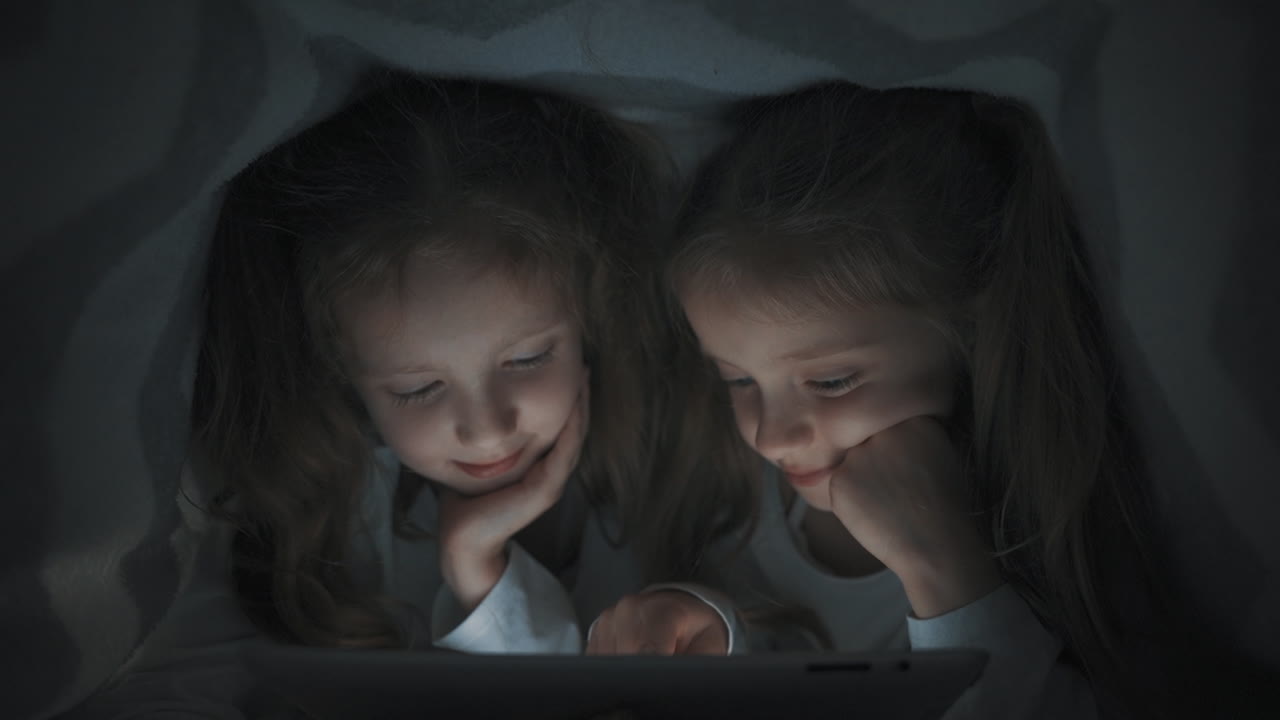 dos niñas usando una tableta escondida en la cama por la noche