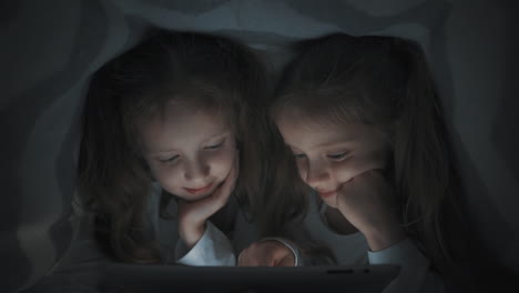 two little girls using a tablet hiding in bed at night