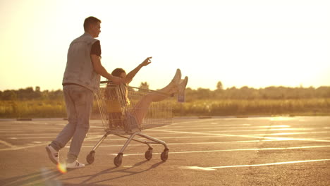 Back-view.-Cheerful-young-couple-in-love-man-and-woman-laughing-and-having-fun-while-riding-carts-in-supermarket-parking-in-slow-motion-at-sunset.