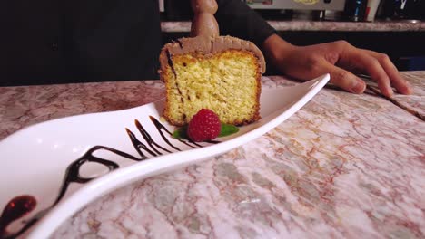 Camarero-Barista-Masculino-Latino-Decorando-Un-Pastel-De-Chocolate-En-Un-Plato-Blanco-Con-Una-Frambuesa-En-Hojas-De-Menta-Verde-En-Un-Restaurante-Cafetería-En-México-América-Latina