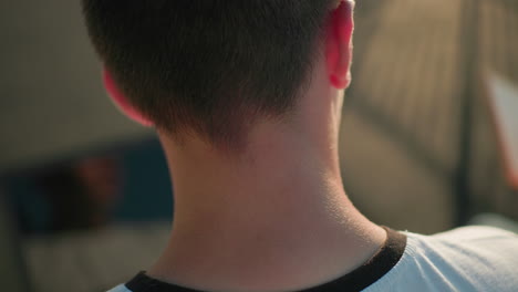 close-up of man's head focusing on neck and ear while glancing sideways at a tablet with his reflection visible on screen, partial blurred background features object and soft lighting