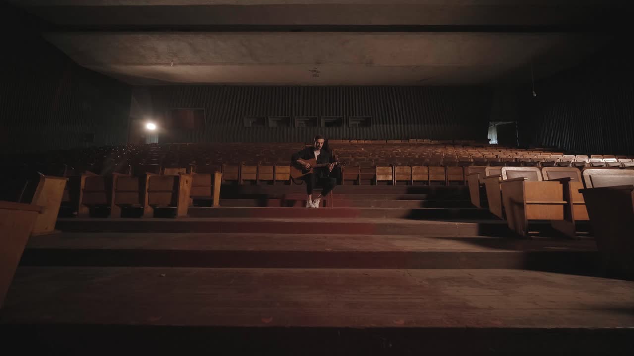 un chico guapo toca una guitarra acústica en un cine abandonado. el músico canta una canción y acompaña en la guitarra
