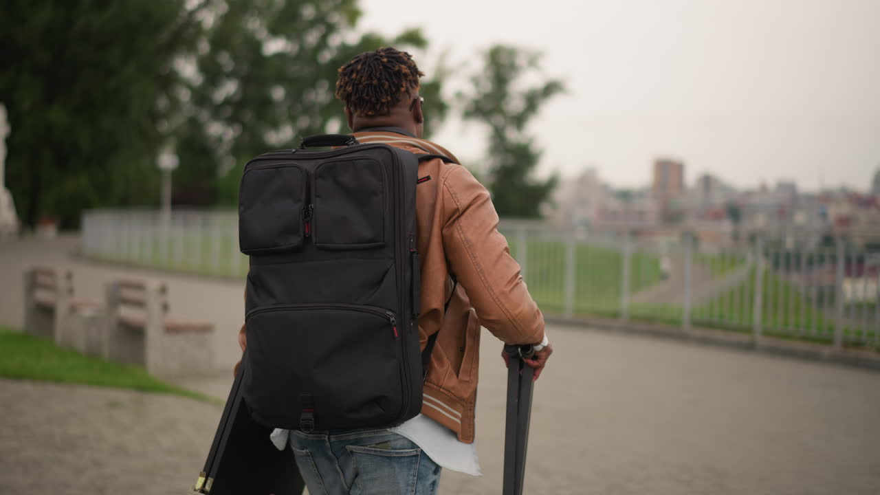 black street musician walking with backpack, leather jacket, carrying tripod and board along riverside promenade, distant city skyline, overcast afternoon, solitary mood, preparing for busking