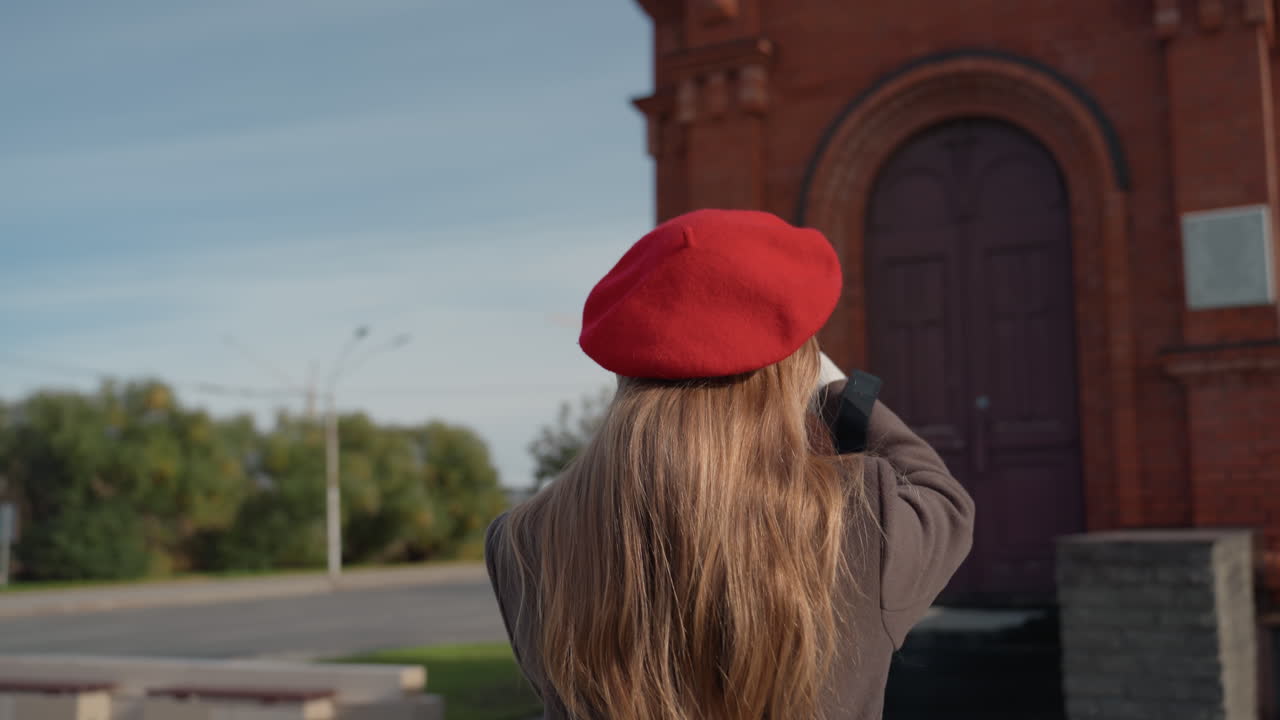 Mujer caucásica fotografiando la cúpula ornamentada de una iglesia, luciendo una boina roja, cabello largo y abrigo marrón. Primer plano de un estudio arquitectónico, con detalles de una cruz dorada y la simetría del campanario; una fotógrafa de arquitectura explorando el patrimonio.