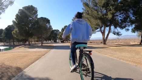 cute african american teenage girl cycling along a park trail on a sunny, windy day - follow view