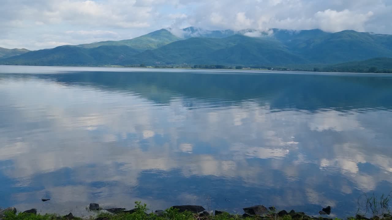 Waters of Kerkini Reservoir in northern Greece reflect surrounding mountains and cloudy sky on a crisp spring day, capturing the serene beauty of the landscape