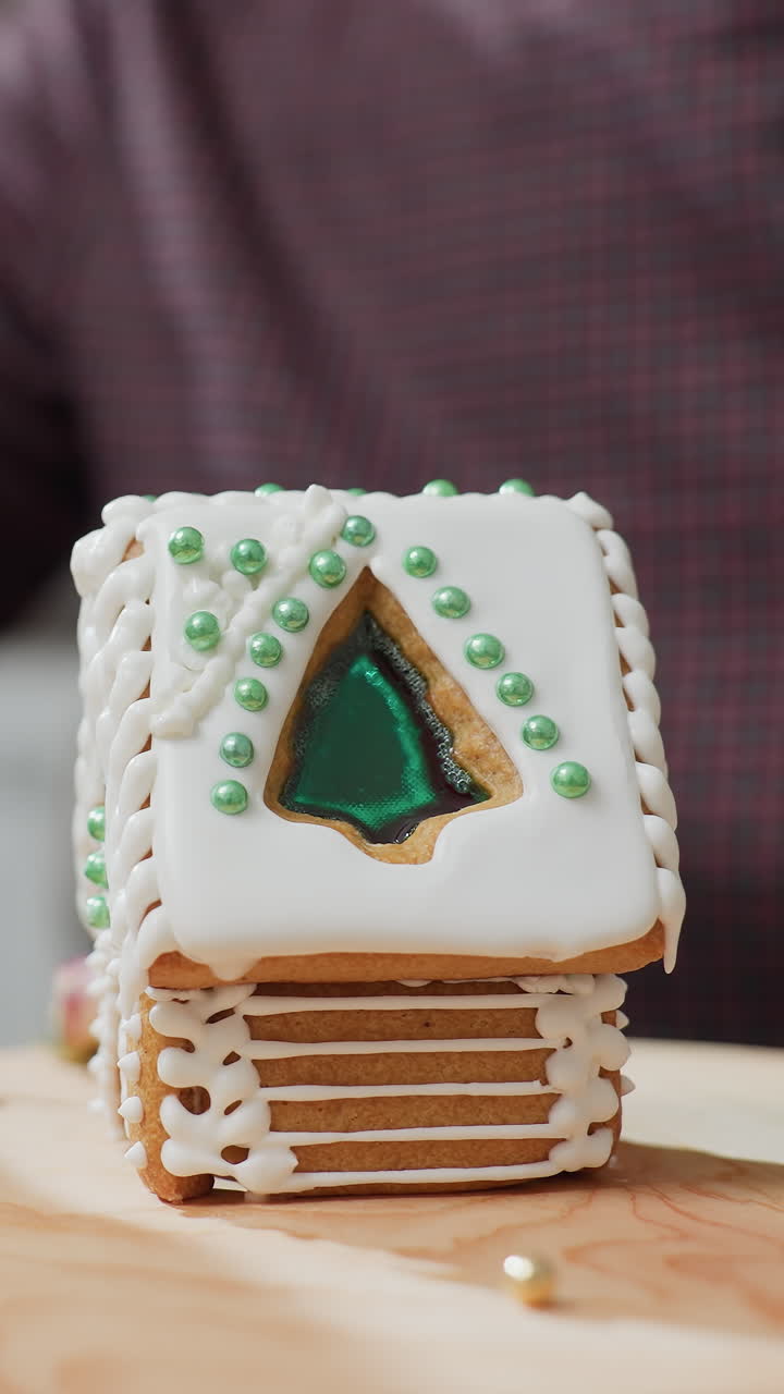 mujer alcanzando para recoger cuentas de oro rodando en una bandeja de madera mientras decora la casa de pan de jengibre, diseño de glaseado intrincado con cuentas verdes, flores secas y luz de vela borrosa en el fondo