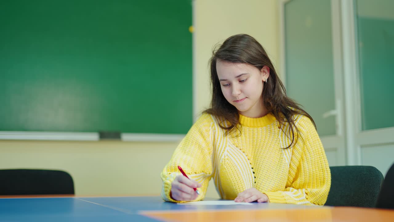 Thoughtful school girl writing at school. Portrait of a beautiful female student sitting at the desk and studying on the classroom background. Education at school.