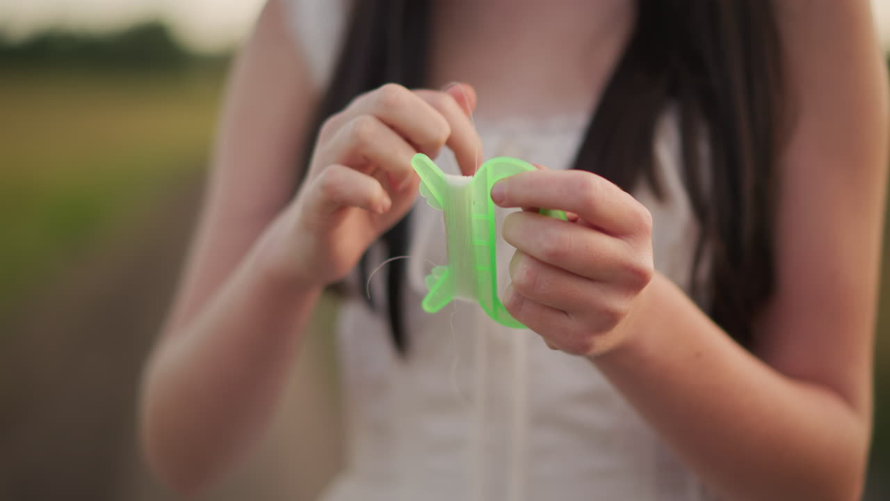 Teenage girl coiling bright kite, Vibrant teenager gently tangling neon kite string during sunset, Young girl meticulously winding luminous kite cord as sunset paints sky in vibrant hues