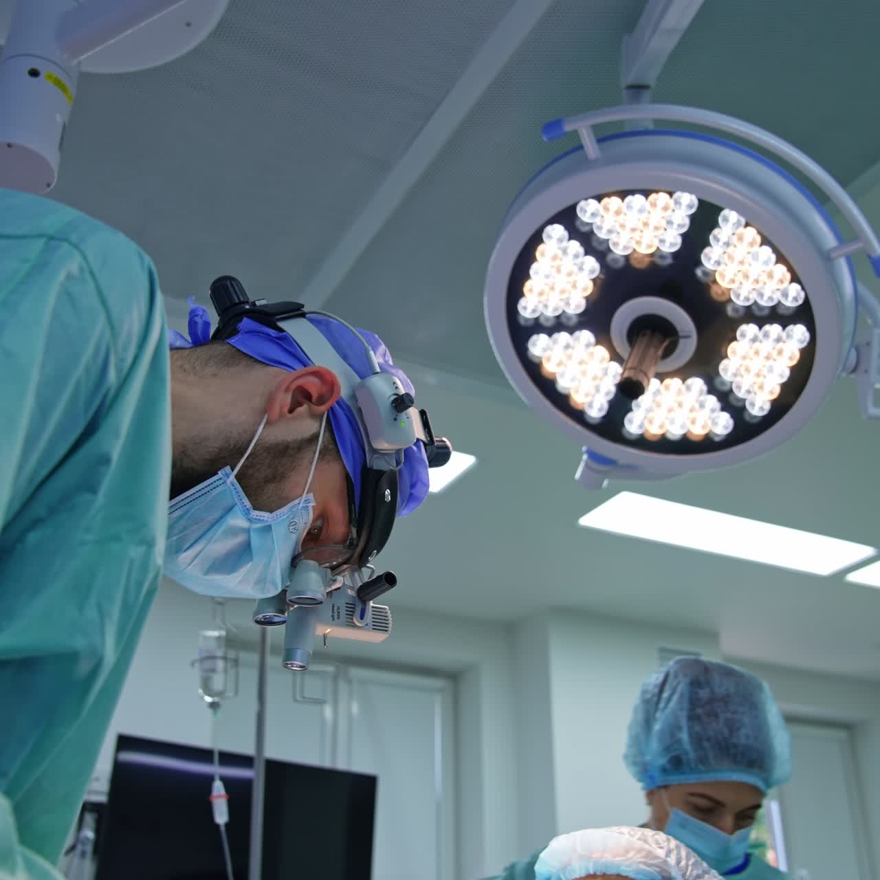 Cirujano en quirófano trabajando con gafas de protección bajo una gran lámpara redonda. La enfermera aparece al fondo.