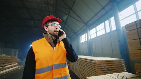 Engineer-in-a-helmet-walks-through-a-warehouse-of-building-materials-and-talks-on-the-phone