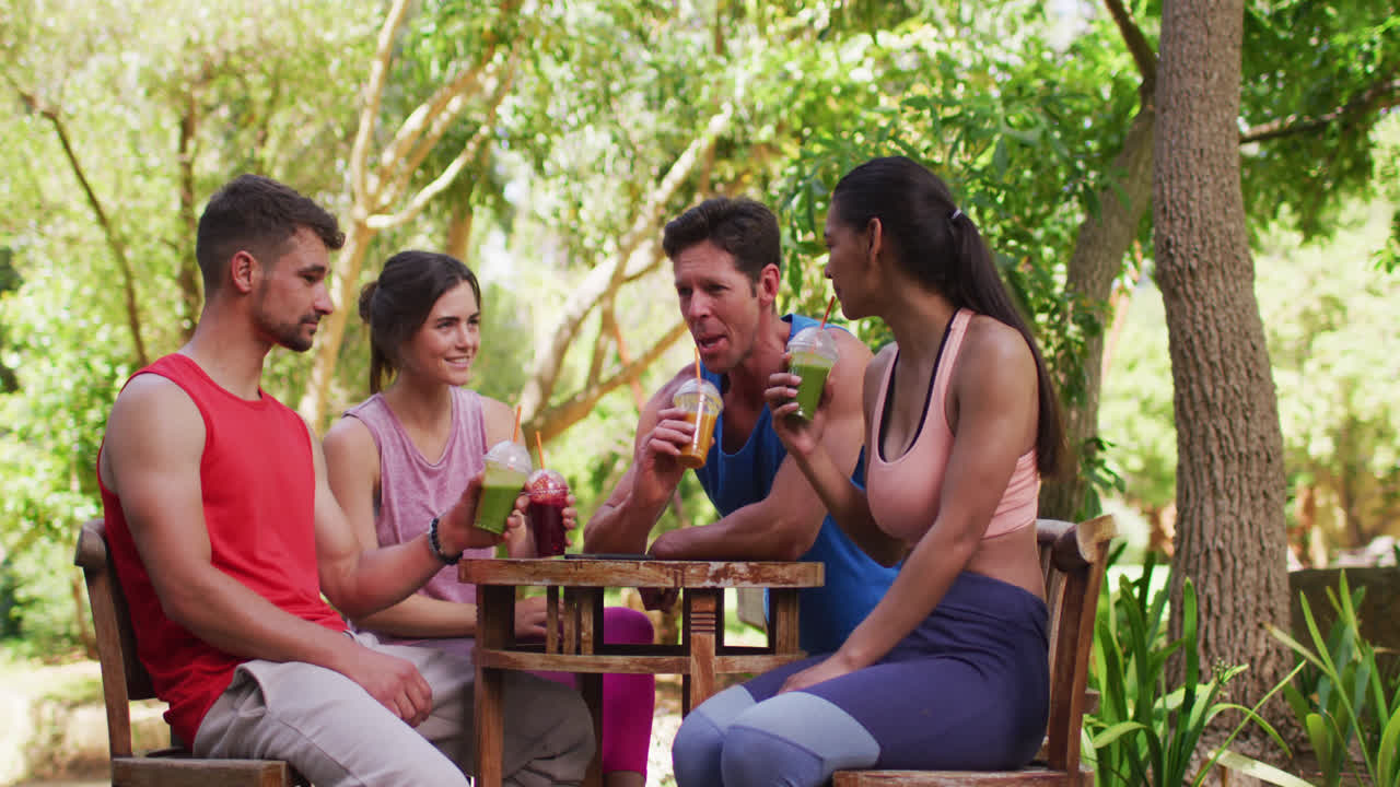 grupo diverso sonriente sentado en la mesa con bebidas saludables, hablando después de yoga en el parque soleado