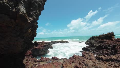 Pan-right-handheld-action-camera-shot-of-small-waves-crashing-into-exposed-coral-rocks-during-low-tide-at-the-famous-tropical-Tambaba-beach-in-Conde,-Paraiba-Brazil-near-Joao-Pessoa-on-a-summer-day