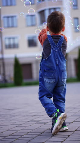 caucasian toddler wearing jeans romper runs outdoors. adorable baby runs catching the soap bubbles. vertical video.