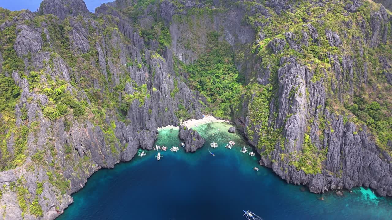 Drone view over Secret Lagoon and Jiji's Beach on Miniloc Island, Philippines, with clear waters, karst cliffs, forested terrain, white sand, and traditional boats floating in a peaceful cove