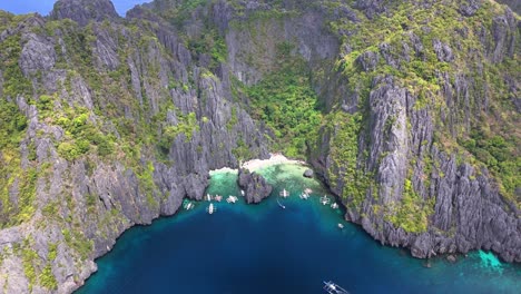 drone view over secret lagoon and jiji's beach on miniloc island, philippines, with clear waters, karst cliffs, forested terrain, white sand, and traditional boats floating in a peaceful cove