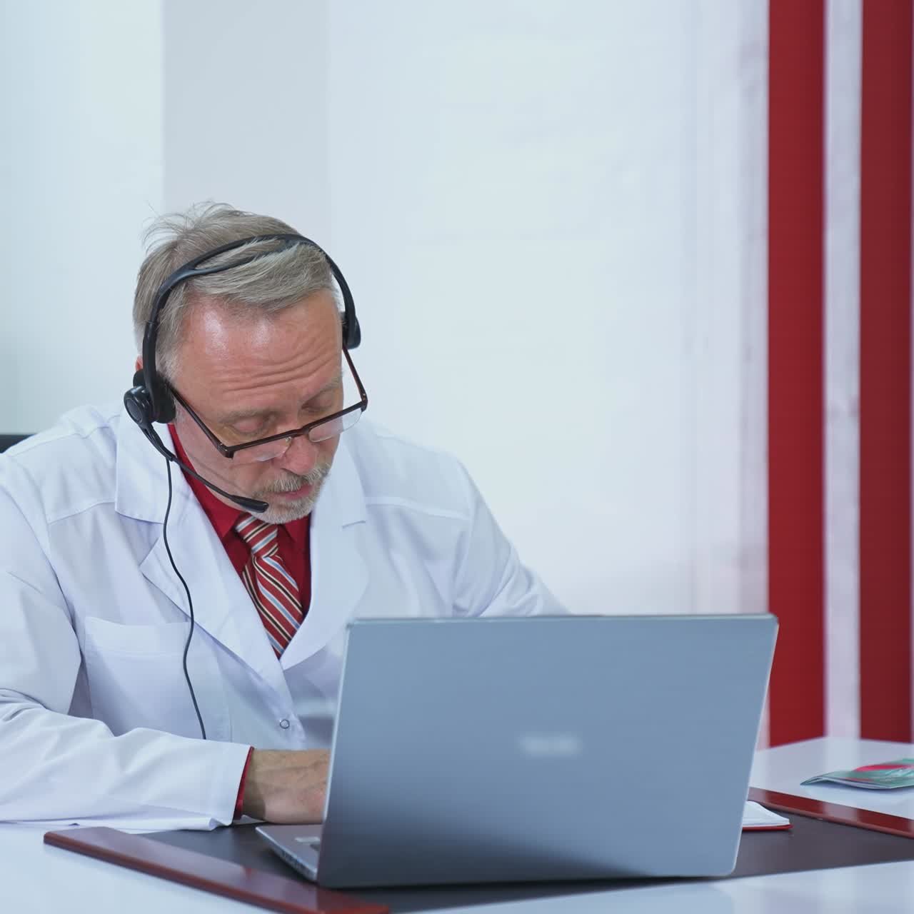 Male physician talking to distant patient. Senior doctor in headset listens to a patient using video chat on a laptop in medical cabinet