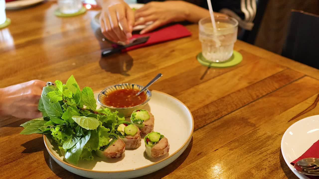 Hands interact with a plate of Vietnamese food on a wooden table, capturing a casual dining moment in natural lighting