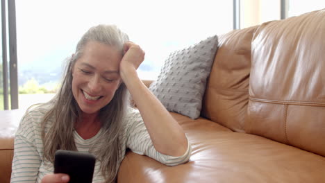 Una-Mujer-Caucásica-Con-Pelo-Gris-Está-Sentada-En-El-Sofá,-Mirando-Un-Teléfono-Inteligente