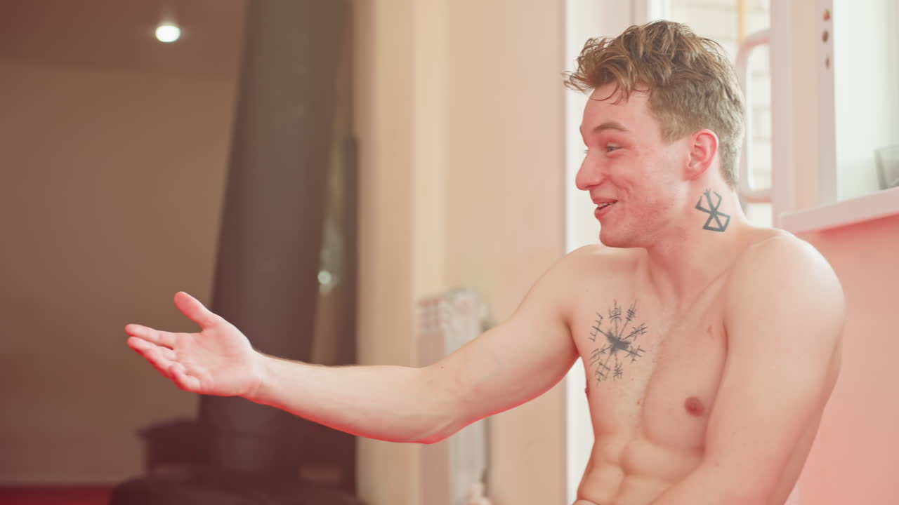 Athlete sitting shirtless inside gym after workout, smiling and looking aside with visible tattoos on chest and neck, showing masculinity, energy, and positive attitude in training environment