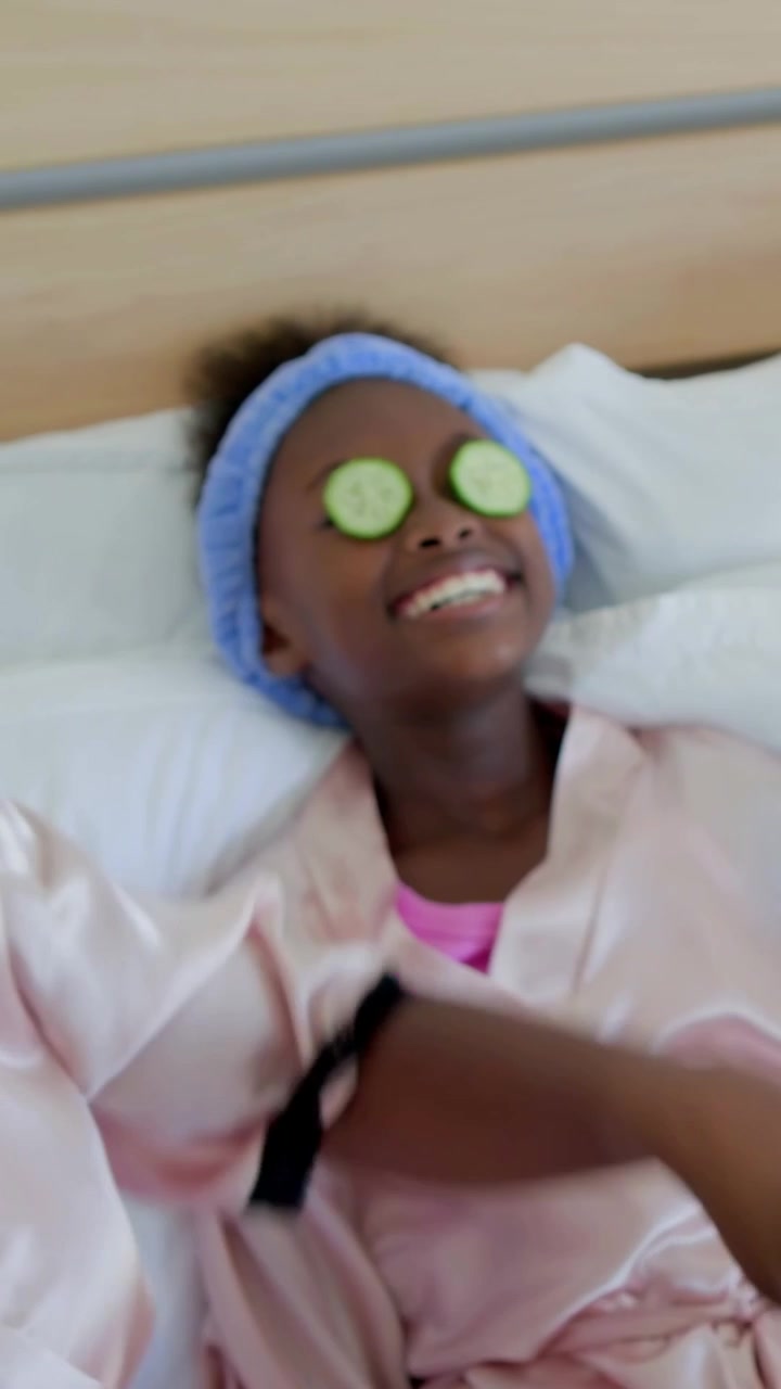 Vertical video: Grabbing bowl, mom in pink robes placing cucumber slices on girls' eyes at home spa