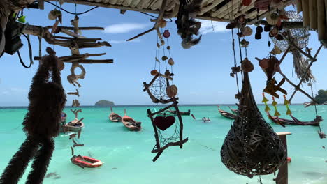 Una-Toma-De-Múltiples-Campanas-De-Viento-Colgando-Y-Mirando-Hacia-El-Océano-Con-Botes-De-Cola-Larga-De-Tailandia-Y-Cielo-Azul-En-El-Fondo