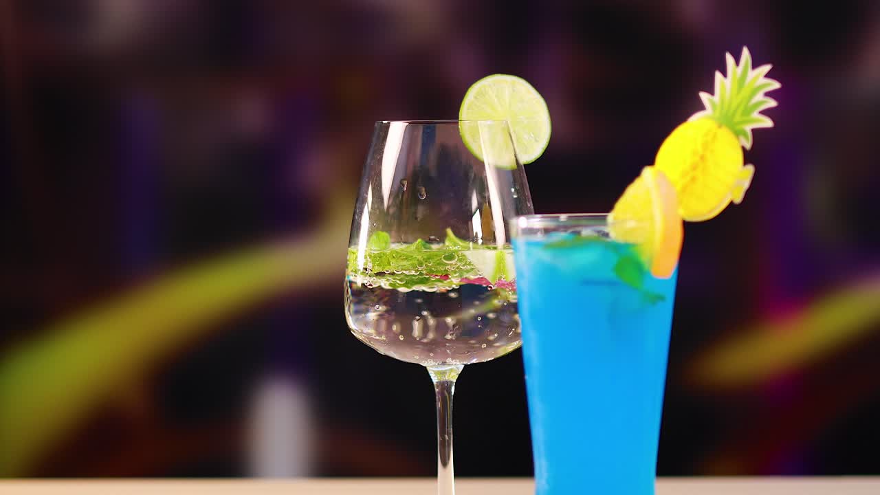 A hand presents a red cocktail alongside blue and clear drinks, set against a lively, colorful bar background